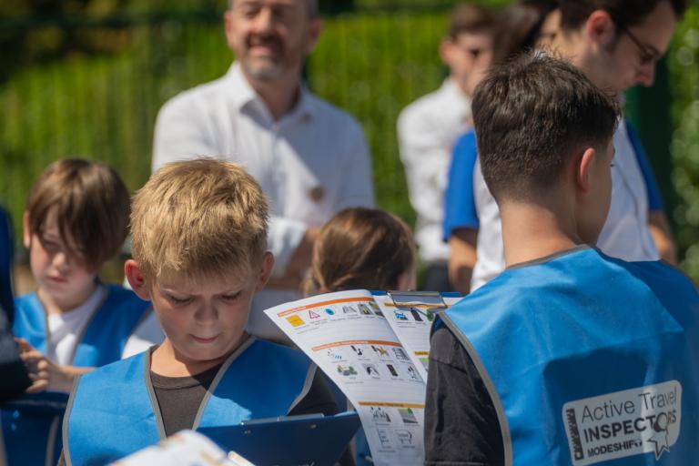 Children wearing blue high visibility vests look at print outs of an inspection toolkit, which includes different street features to tick off. Chris Boardman can be seen in the background.