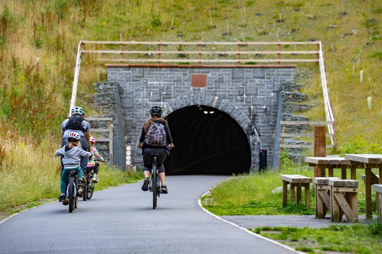 Two adults and two children cycle along a tarmac path towards a tunnel in a hillside. The tunnel is lit inside. Next to the path is a wooden picnic bench with seating.