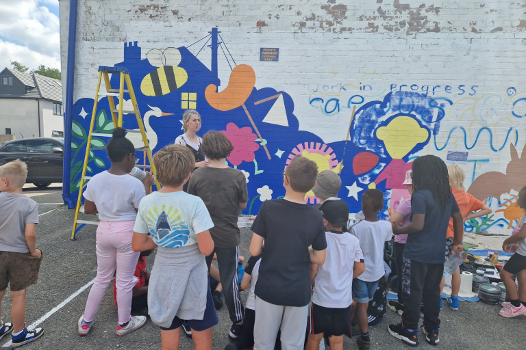 Children taking part in a mural painting exercise. There is a brightly coloured mural painted on an outside wall behind them. The mural includes shapes such as a bee and a house.