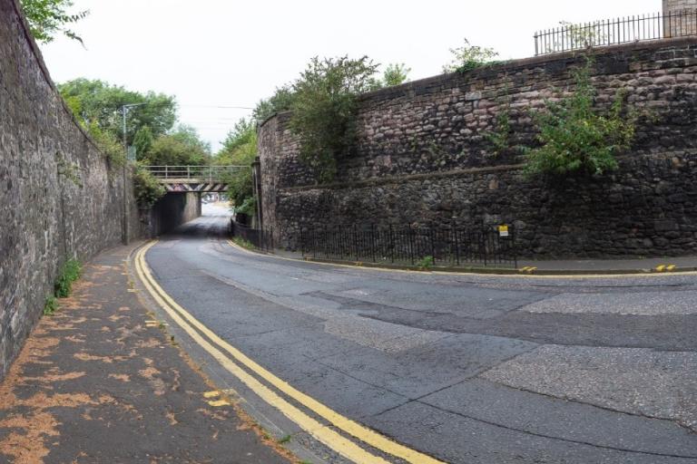 A road passing under a bridge. There are double yellow lines by the kerb all along the road. The pavement on each side of the road is bordered by stone walls.