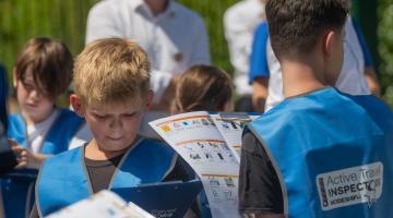 Children wearing blue high visibility vests look at print outs of an inspection toolkit, which includes different street features to tick off. Chris Boardman can be seen in the background.