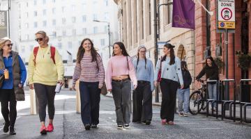 A group of women walk along a pavement in an urban area.